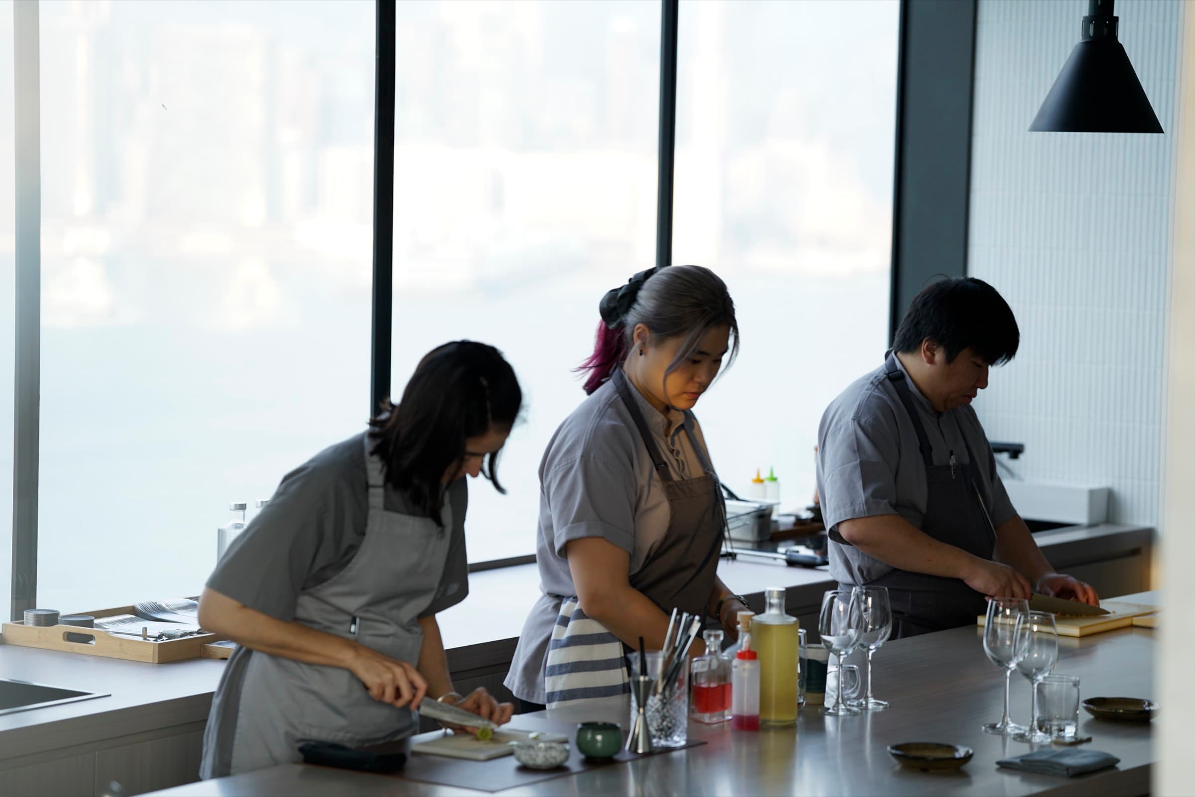 Chef Chevalier Yau and the æra kitchen team preparing dishes at the pass, with Victoria Harbour visible through the window